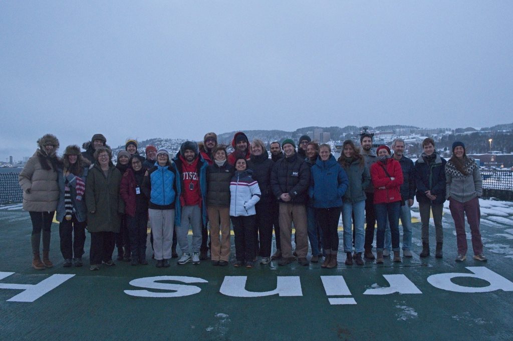 Group photo of researchers and artists participating in the EXTREME25 expedition on the deck of the RV Kronprins Haakon, dressed in winter clothing against a snowy backdrop.