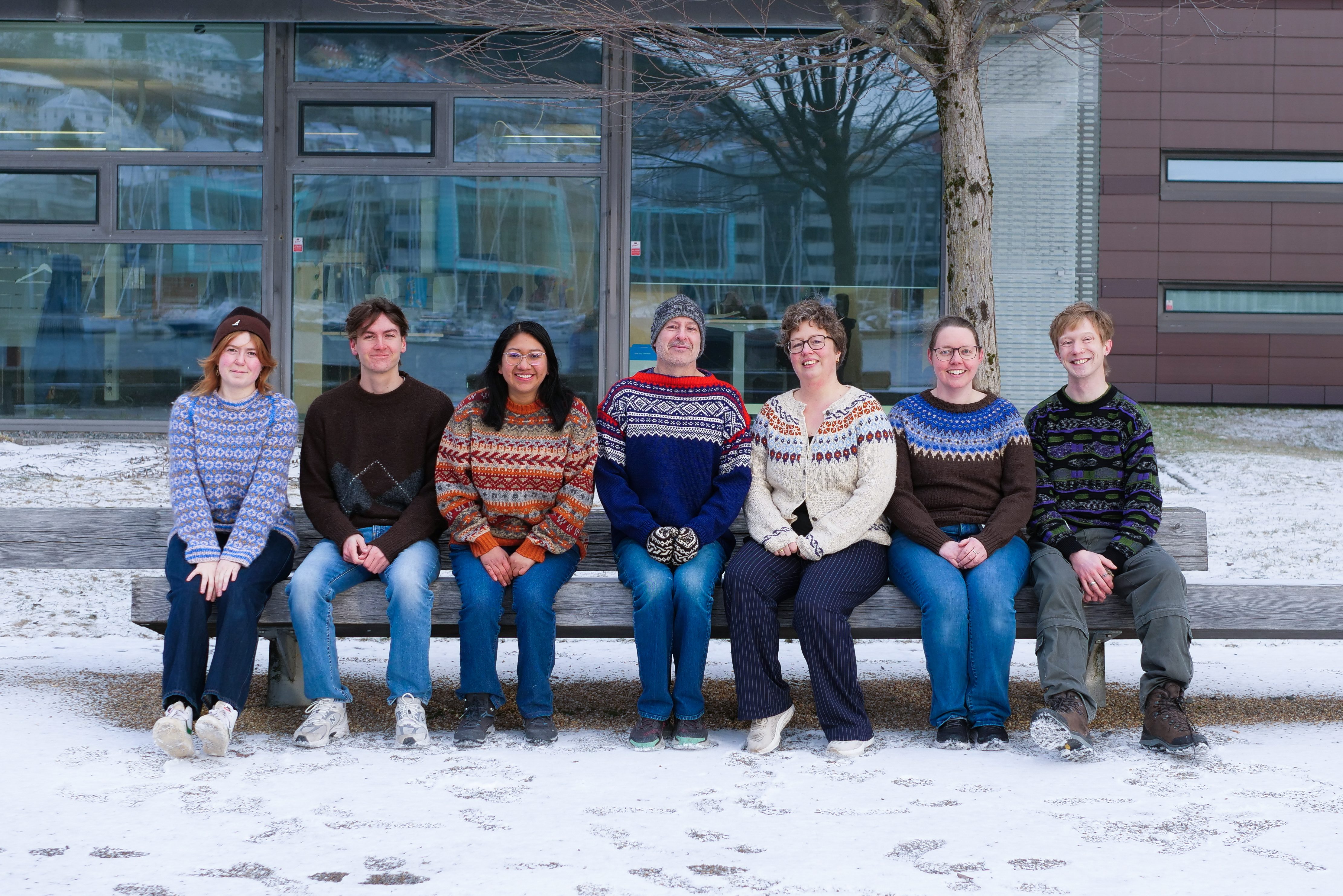 Researchers sitting on a bench in the snow in colorful knitted sweaters.